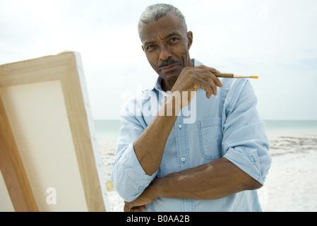 Senior man standing in front of canvas at the beach, holding paint brush, smiling at camera Stock Photo