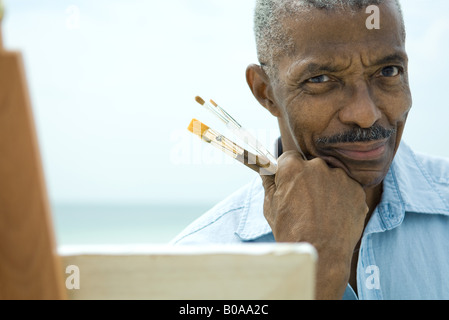 Senior man with hand under chin, holding paint brushes, smiling at camera, close-up Stock Photo