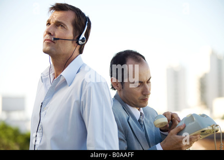 Two men standing back to back, one using headset, the other holding landline phone Stock Photo