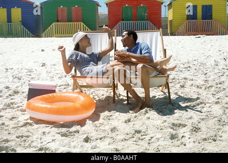 Couple sitting in lounge chairs at the beach, smiling, woman's legs on man's lap Stock Photo