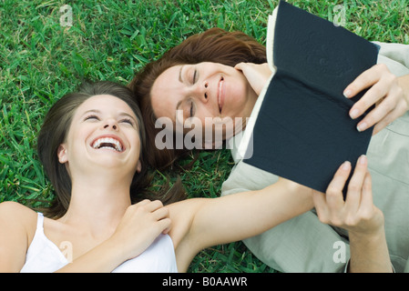 Mother and teen daughter lying together in grass, laughing, woman holding book Stock Photo