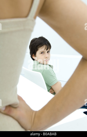 Boy sulking, looking over shoulder at mother in foreground with hand on hip Stock Photo