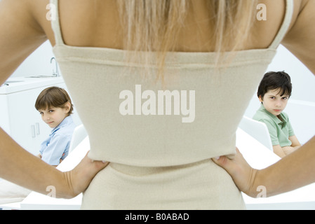 Mother standing with hands on hips, rear view, boys sulking in background, cropped view Stock Photo