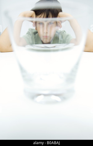 Boy sulking at table, looking at camera through water glass Stock Photo