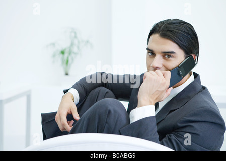 Young businessman sitting with knees up, using cell phone, looking at camera Stock Photo