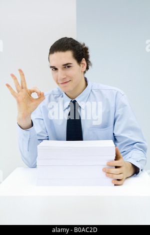 Young professional sitting behind stack of papers, making OK hand gesture, portrait Stock Photo