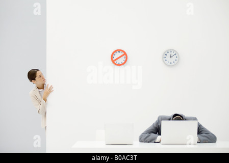 Female office worker peeking around corner at clocks on wall, colleague sitting in front of laptop with head down Stock Photo