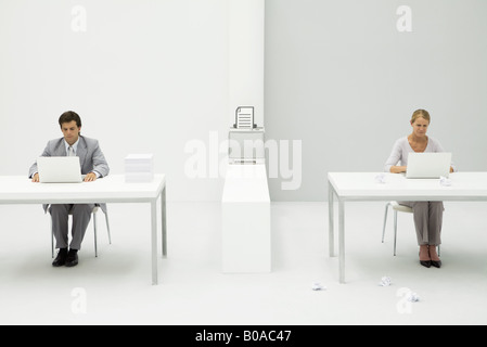 Colleagues in office using laptop computers, printer between them with graphic of document, paper balls scattered on the floor Stock Photo