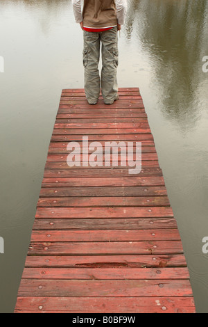 Teenage boy standing at the end of a wooden pier jetty on a lake as it starts to rain Stock Photo