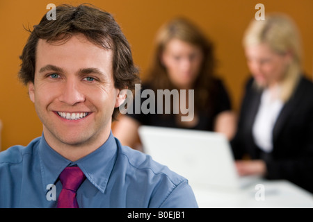 A happy young male executive in focus in the foreground while his female colleagues work on a laptop behind him Stock Photo
