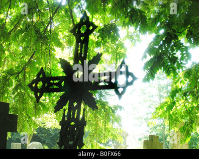 silhouette of wrought iron grave yard cross in backlighting against green sunlit leaves Stock Photo