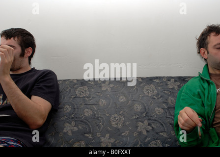 Gap between two young white males sitting on a Grey sofa both are reading newspapers and one has a joint, only sides of faces. Stock Photo