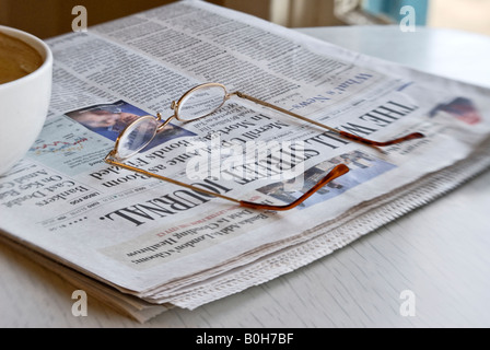 a cup of coffee, reading glasses, and newspaper sit on a shiny white tabletop Stock Photo