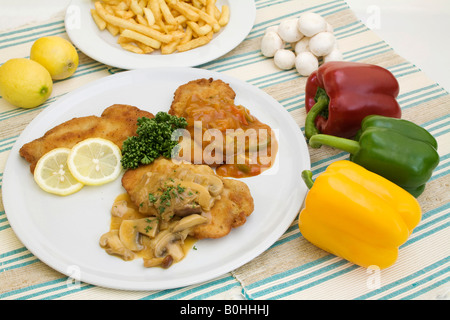 Wiener schnitzel served on a white plate surrounded by a green, red and yellow peppers, mushrooms, lemons and a bowl of french  Stock Photo