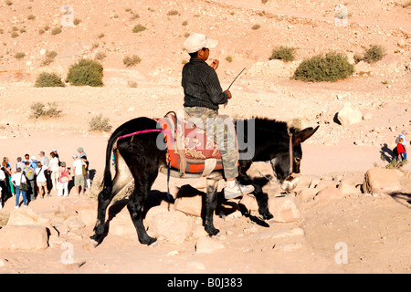 Bedouin Arab boy in baseball cap on donkey by salmon pink and red sandstone hills of Petra in Jordan by the Colonnaded Street Stock Photo