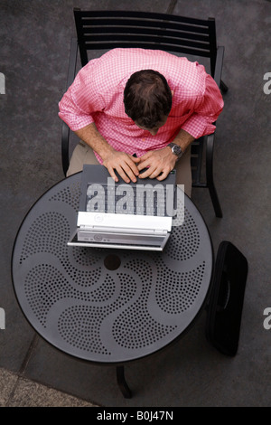 A young man sits at an outdoor cafe, working on his laptop computer. Stock Photo