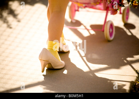 young girl walking in mom's shoes, pushing toy baby stroller, color, close up shoes and leg Stock Photo