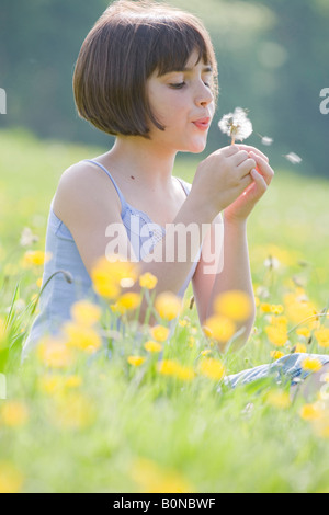young female child sitting in field of buttercups blowing a dandelion Stock Photo