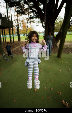 Four year old girl on a swing, mother pushing and brother in background Stock Photo
