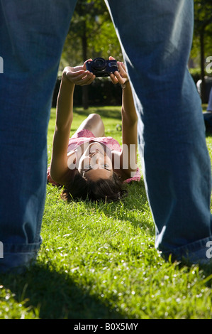 A young woman lying on grass and taking a photograph of a man standing Stock Photo