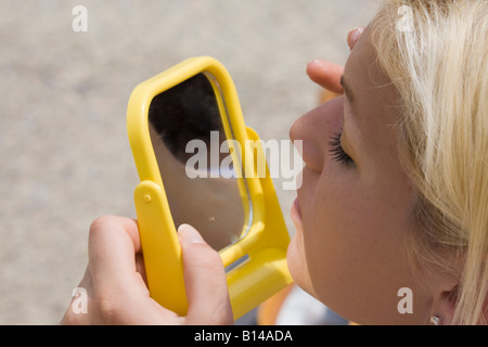 Europe Attractive young blonde haired woman using yellow hand-held mirror applying eye face makeup Stock Photo