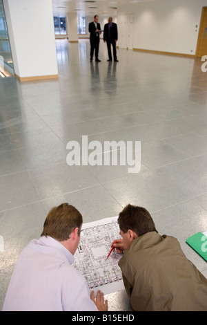 Two engineers inspect some plans as two men in suits read a clipboard in the background. Stock Photo