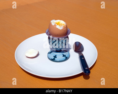 Brown soft boiled egg with top removed in blue green egg cup with teaspoon on white plate set on sunlight light brown table Stock Photo