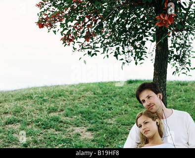 Couple sitting under tree, sharing earphones Stock Photo
