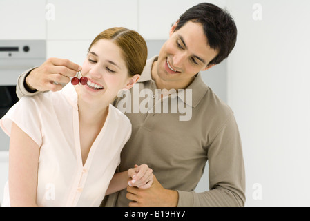 Young couple in kitchen, man holding cherries in front of woman's mouth Stock Photo