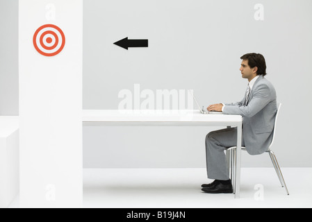 Man sitting at desk, using laptop computer, arrow on wall pointing at bull's-eye Stock Photo