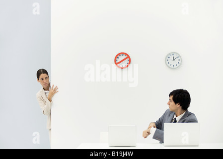 Woman peeking around corner as boss points at wristwatch, clocks on wall in background Stock Photo