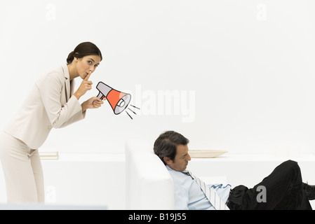 Man sleeping in chair, woman behind him holding megaphone, looking at camera, finger over lips Stock Photo