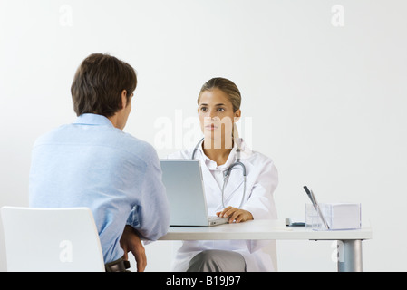 Female doctor sitting across from male patient at desk, using laptop computer Stock Photo