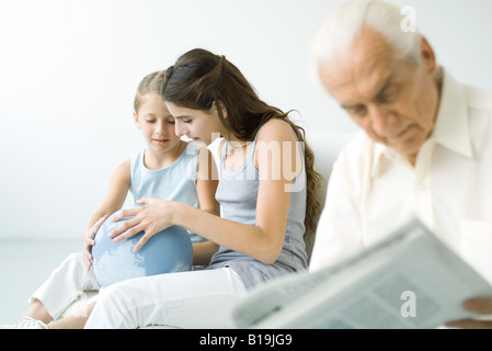 Preteen girl and younger sister looking at globe together, senior man reading newspaper in foreground Stock Photo