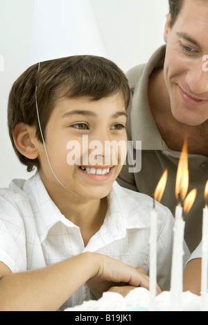 Boy next to birthday cake with lit candles, wearing party hat, smiling Stock Photo