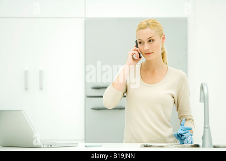 Young woman standing in kitchen, holding rubber gloves, using cell phone Stock Photo