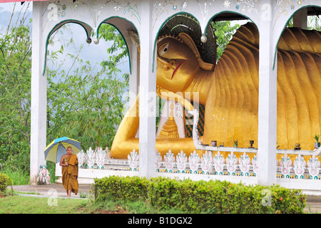 A senior Buddhist monk with umbrella in front of reclining Buddha. Sangkhla Buri, Thailand. Stock Photo