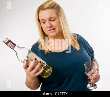 overweight blonde young woman drinking white wine and looking at the bottle in a worried concerned way, in two minds Stock Photo
