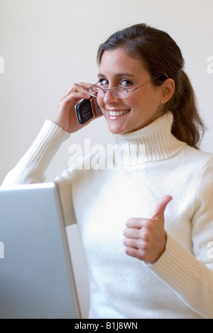 young pretty woman with white turtleneck sweater and glasses, working with a notebook, phoning, making thumb up Stock Photo