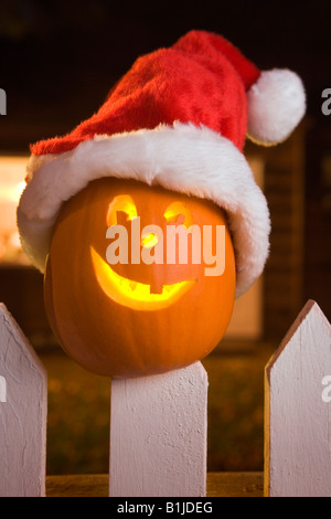 Jack-O-Lantern face wearing a santa hat stuck on top of a fence post of white picktet fence at twilight during Fall Stock Photo