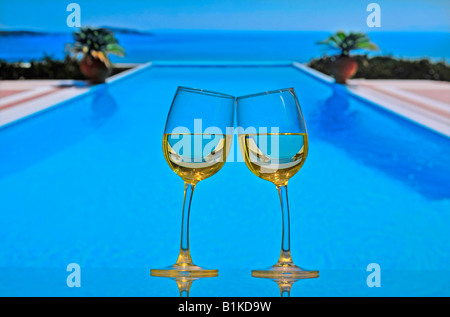 Quirky holiday wine celebration Two white wine glasses on table lean towards each other and 'chink', with infinity swimming pool and sea in background Stock Photo