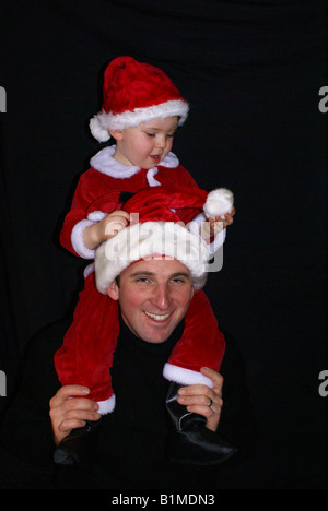 small boy in Santa suit  on Dad's shoulders examining white tassel on Dad's Santa hat Stock Photo
