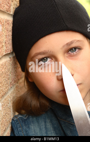 Young teenager girl with sharp knife denim jacket and black beany hat in urban street alley with brick wall POSED BY MODEL Stock Photo