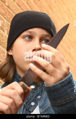 Young teenager girl with sharp knife denim jacket and black beany hat in urban street alley with brick wall POSED BY MODEL Stock Photo