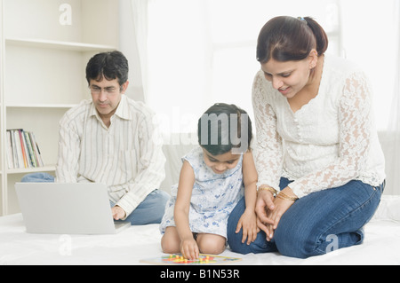 Mid adult woman and her daughter playing a board game with a mid adult man using a laptop behind them Stock Photo