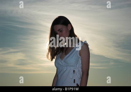 Silhouette of a young woman wearing white dress standing outdoors at dusk sun shining behind head bowed