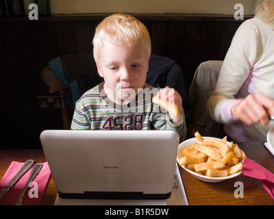 Young child / boy / kid watches / watching a TV film on a portable DVD player while eating chips. UK Stock Photo