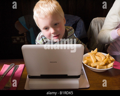 Young child / boy / kid watches / watching a TV film on a portable DVD player while eating chips. UK. Stock Photo