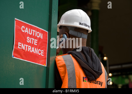 A black man wearing a protective helmet and fluorescent jacket speaks on his mobile phone at a London construction site entrance Stock Photo