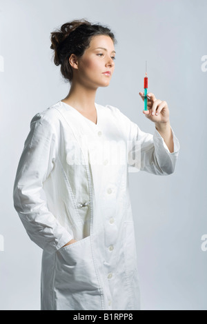 Young woman wearing a white laboratory smock looking concentrated while holding a syringe filled with red liquid in her hand Stock Photo